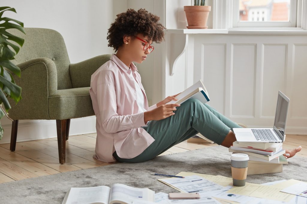 Sideways shot of serious fashionable woman wears optical glasses, checks written article, reads book