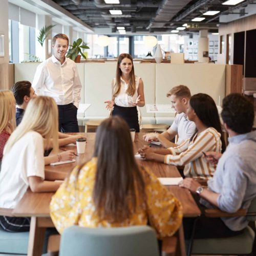 Businessman And Businesswoman Addressing Group Of Young Candidates Sitting Around Table And Collaborating On Task At Graduate Recruitment Assessment Day Businessman And Businesswoman Addressing Group Of Young Candidates Sitting Around Table And Collaborating On Task At Graduate Recruitment Assessment Day