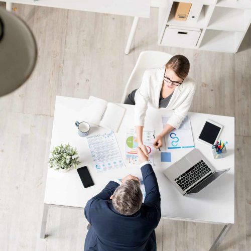 Businessman concluding agreement with audit specialist Top view of confident businessman in suit sitting at table with charts documents and making handshake with audit specialist while concluding agreement at meeting